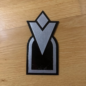 May include: A black and silver 3D printed emblem with a geometric design. The emblem features a diamond shape at the top, a V-shaped design in the center, and a rounded arch shape at the bottom.