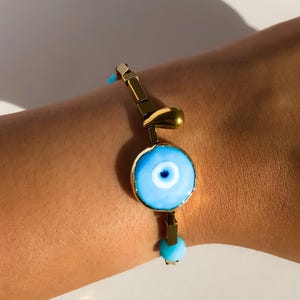 May include: A gold bracelet with a blue evil eye charm and a teardrop-shaped pendant. The bracelet is made of small gold squares and beads.