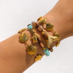 May include: A set of four gold bracelets with a sea-themed design. The bracelets feature seashells, a turtle, and a mermaid tail charm. The bracelets are made of gold-toned metal and have a brown and blue beaded design.