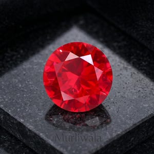 May include: A round, faceted, vibrant red gemstone with a brilliant cut, displaying multiple triangular and kite-shaped facets that reflect light. The gemstone is positioned on a dark, square, polished stone base with the text Murliwala visible.