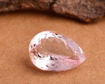Puede incluir: Una piedra preciosa de color rosa pálido en forma de pera con facetas, descansando sobre una superficie marrón.