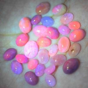 May include: A collection of oval-shaped gemstones in shades of pink, purple, and orange. The stones are smooth and polished, with a glowing, iridescent quality. The gemstones are scattered across a light-colored surface.