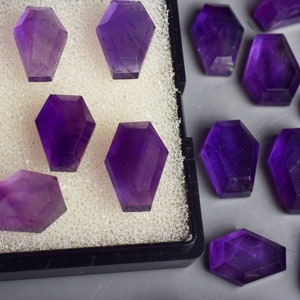 Puede incluir: Una colección de piedras preciosas de amatista púrpura, talladas en forma de ataúd, expuestas en una caja de terciopelo negro y esparcidas sobre una superficie gris.