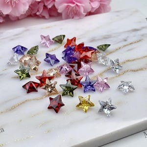 Puede incluir: Una colección de cristales en forma de estrella facetados y coloridos en varios colores, incluyendo rojo, rosa, morado, verde y transparente. Los cristales están dispersos sobre una superficie de mármol blanco y gris, con flores rosas en el fondo.