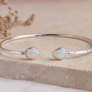 May include: A silver cuff bracelet featuring a hammered texture along the band and two teardrop-shaped opal stones at the open ends. The iridescent opals display flashes of blue and green colour, set within smooth silver bezels on the metal frame.