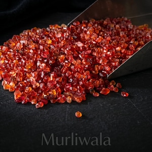 May include: A pile of small, translucent, deep red garnet gemstone chips being poured from a silver metal scoop. The faceted stones exhibit varying shades of crimson and orange, and the word Murliwala is printed at the bottom of the frame.