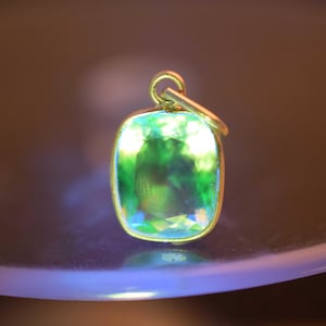 Könnte beinhalten: Ein goldener Anhänger mit einem rechteckigen, smaragdgeschliffenen Edelstein. Der Stein hat eine lebendige grün-blaue Farbe, gefasst in einer goldenen Lünette. Der Anhänger hängt an einer kleinen Goldschlaufe.