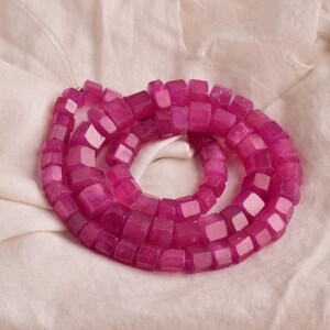 Può includere: Una collana di perline sfaccettate di colore magenta. Le perline sono a forma di cubo con molteplici sfaccettature, infilate su un filo sottile e trasparente. Le perline sono disposte in un motivo circolare su una superficie di tessuto bianco.