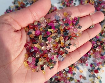 Könnte beinhalten: Eine Handvoll bunter, roher Edelsteine in verschiedenen Größen und Formen. Die Edelsteine zeigen eine Mischung aus Farben, darunter Rosa, Gelb, Blau, Grün und Orange. Die Edelsteine sind auf einer weißen Oberfläche verstreut.