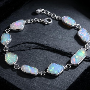 May include: A silver-toned chain bracelet featuring seven irregular, bezel-set opal stones that display iridescent flashes of blue, green, and pink. The bracelet includes a lobster claw clasp and an adjustable extender chain with a small round bead.