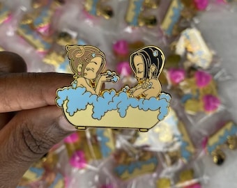 May include: A gold-colored enamel pin featuring two cartoon figures in a yellow bathtub with blue bubbles. The figures have brown and black hair. Many similar pins are visible in the background, packaged in clear plastic.