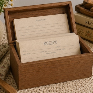 Puede incluir: Una caja de recetas rectangular de madera con tapa abatible, que contiene una pila de tarjetas de recetas de color tostado claro. Las tarjetas presentan texto impreso que incluye las palabras RECIPE, Directions, Title, Prep Time, Cook Time, Serves, Preheat Oven e Ingredients.