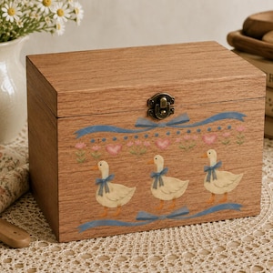 Puede incluir: Una caja rectangular de madera con tapa abatible y cierre met&aacute;lico. La parte frontal presenta un dise&ntilde;o impreso de tres patos blancos con lazos azules, rodeados de cintas azules, corazones rosas y peque&ntilde;os detalles florales verdes.