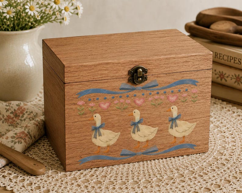 Puede incluir: Una caja rectangular de madera con tapa abatible y cierre met&aacute;lico. La parte frontal presenta un dise&ntilde;o impreso de tres patos blancos con lazos azules, rodeados de cintas azules, corazones rosas y peque&ntilde;os detalles florales verdes.