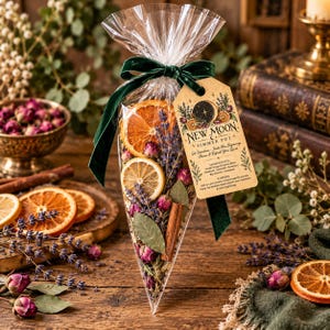 May include: A clear cone-shaped bag filled with dried orange slices, lavender sprigs, pink rosebuds, bay leaves, and cinnamon sticks. A dark green velvet ribbon ties the top, and a tan tag reads New Moon Simmer Pot with listed ingredients.