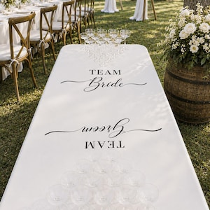 May include: A white rectangular table cover featuring black text that reads Team Bride and Team Groom, arranged for a game. The table is set with two triangular formations of clear stemmed glasses on opposite ends of the surface.