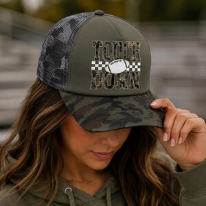 May include: An olive green trucker hat with a camouflage print brim and mesh back. The front panel features the text TOUCH DOWN in a leopard print pattern, separated by a white football graphic and a black and white checkered horizontal stripe.