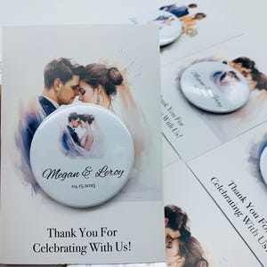 May include: A white wedding favor button with a watercolor image of a couple, the names "Megan & Leroy" and the date "04.15.2025". The button is on a card that says "Thank You For Celebrating With Us!"
