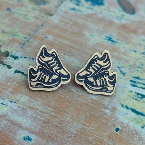 May include: Wooden stud earrings featuring a navy blue and tan sneaker design. The earrings are in the shape of two stacked sneakers. The earrings are on a light blue and tan wooden surface.