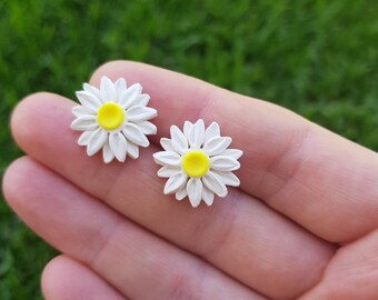 May include: A pair of white daisy earrings with yellow centers. The earrings are shown on a hand against a green background.