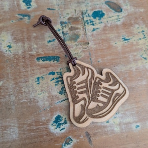 Puede incluir: Un llavero de madera con un diseño de dos zapatillas deportivas. Las zapatillas están grabadas en la madera, mostrando los detalles del diseño del calzado. Una cuerda de cuero marrón está unida al llavero.