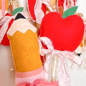 May include: A felt pencil and red apple decorations with pink ribbons. The pencil is yellow with a white tip and pink band. The apple has a green leaf and brown stem. The decorations are on wooden sticks.