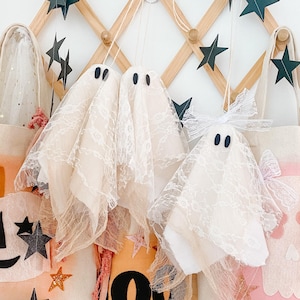 May include: Three hanging ghost ornaments made of white lace fabric with black felt eyes, suspended by strings. Below them are three canvas tote bags featuring painted designs, including a pink skull, an orange bag with the text BOO, and stars.
