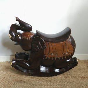 May include: A dark brown wooden rocking elephant with a smooth, polished finish. The elephant features a carved saddle in a lighter tan shade with decorative etched details, a raised trunk, and curved rockers at the base for a gentle rocking motion.