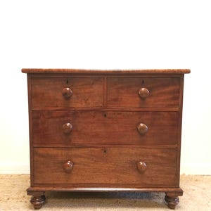 May include: A dark brown wooden chest of drawers with four drawers and round knobs. The chest has a classic design and sits on four small, rounded feet. The background is a plain white wall. The chest is made of wood.