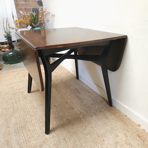 May include: A dark wood drop-leaf table with black legs. The table has a rectangular top with rounded corners and a dark finish. The legs are slender and angled, and the table is set on a woven rug.