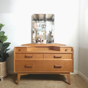 May include: A light brown wooden vanity dresser featuring a rectangular mirror attached to the top. The piece includes two small upper drawers with round knobs and two larger lower drawers with horizontal wooden handles, all supported by four legs.