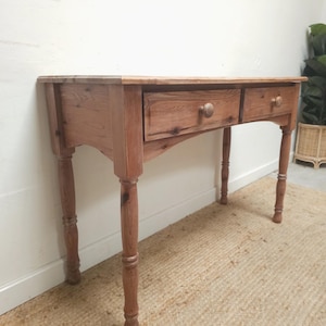 May include: A light brown wooden console table with two drawers and turned legs. The table is made of wood and has a natural finish. The drawers have round wooden knobs. The table is placed against a white wall and sits on a beige rug.