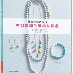 Könnte beinhalten: Ein Buchcover mit dem Titel "Fünf bunte Papierperlen-Schmuckstücke" in japanischen Schriftzeichen. Das Cover zeigt eine Auswahl an Papierperlen-Schmuck, darunter Halsketten, Ohrringe und ein Armband. Die Perlen sind aus weißem, blauem und rosa Papier gefertigt.