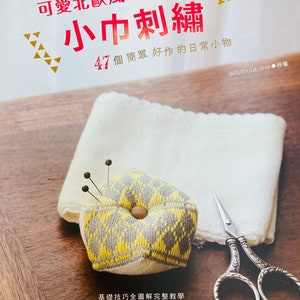 May include: A close-up of a yellow and gray pincushion with a hexagonal shape. The pincushion is filled with pins and is sitting on a white cloth. A pair of silver scissors is also visible in the image. The text on the image reads "可愛北歐風の 小巾刺繡 47個簡單好作的日常小物 基礎技巧全圖解完整教學 初學者一看就會 BOUTIUQE-SHA◆授權".