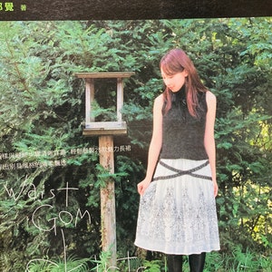 May include: A book cover with a black background and white text that reads "25款優雅飄逸的手作長裙 鬆緊腰裙的浪漫DIY 3覺著". The cover features a woman wearing a black sleeveless shirt, a white skirt with a black floral pattern, and black boots. She is standing in a garden with green trees and bushes.