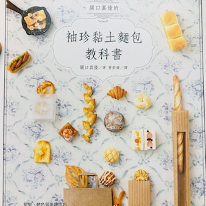 Op de afbeelding: Een boekomslag met een witte achtergrond en een bloemenpatroon. De titel is "Miniature Clay Bread Textbook" in het Japans. De omslag toont verschillende miniatuur broodcreaties van klei, waaronder een stokbrood, een croissant, een pretzel en een brood.