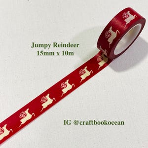 Op de afbeelding: Rode washi tape met een herhalend patroon van crèmekleurige rendieren. De tekst "Jumpy Reindeer 15mm x 10m" is op de tape gedrukt. De Instagram-handle @craftbookocean is ook zichtbaar.