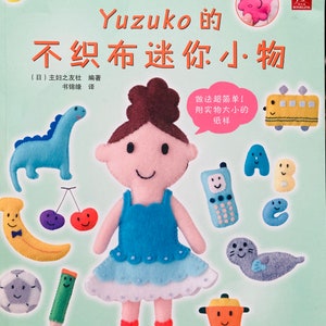 May include: A book cover with a colorful illustration of a girl and various felt toys. The title of the book is "Yuzuko's Felt Miniatures" in Chinese characters. The book cover is green and features a variety of felt toys, including a dinosaur, a banana, a cherry, a soccer ball, a pencil, a phone, a seal, a pot, and letters A, B, and C.