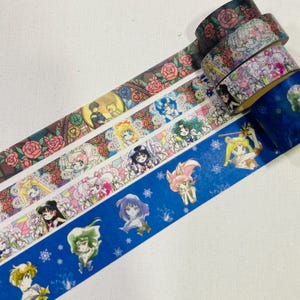 May include: Three rolls of decorative adhesive tape featuring anime-style character illustrations and floral patterns. One roll displays pink roses, another shows various anime figures with flowers, and the third is blue with character portraits.