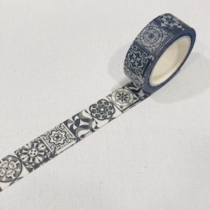 Op de afbeelding: Decoratieve washi-tape rol met een herhalend patroon van grijstinten geometrische tegelontwerpen. De tape is afgerold en toont het patroon. De rol ligt op een witte ondergrond.