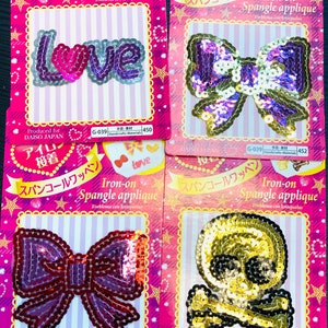 May include: Four iron-on spangle appliques on pink cards. Designs include the word "LOVE", a purple bow, a red bow, and a gold skull with crossbones. Each card has Japanese text and the words "Iron-on Spangle applique".