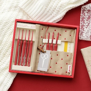 May include: A red box set containing crochet hooks with cushion grips. The set includes several red crochet hooks, a beige fabric case with red polka dots, and a tag with scissors. The box is on a red surface.