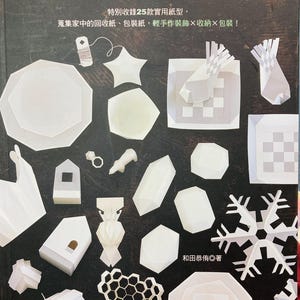 Puede incluir: Una portada de libro con el título "Geometría creativa, juego de papel" en caracteres japoneses. La portada presenta una variedad de diseños de origami de papel blanco, incluyendo un copo de nieve, una casa, una estrella y un panal.