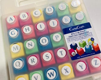 May include: A translucent plastic carrying case containing a set of twenty-six colourful square craft punches, each labelled with a letter of the alphabet from A to Z. A blue label on the case reads CarlaCraft Mini Craft Punch Upper Case Alphabet Set.