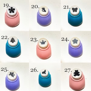 May include: A collection of nine craft paper punches labeled 19 through 27, featuring various shapes including a flower, hand gesture, butterfly, tree, crown, star, angel, seal, and bear, set in pink, purple, and blue plastic housings.