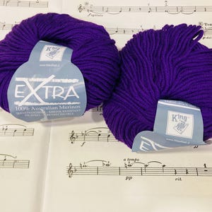 May include: Two round skeins of deep purple yarn, each featuring a light blue paper label. The labels display the text King, EXTRA, and 100% Australian Merinos. The yarn is composed of thick, twisted fibers, presented as a pair of identical units.