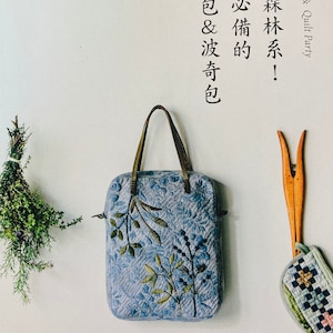 May include: A blue quilted tote bag with a floral design and brown leather handles. The bag is on a white background with a sprig of greenery to the left. The text "愛上森林系! 職人必備的 拼布包&波奇包" is written in black on the right side of the image.