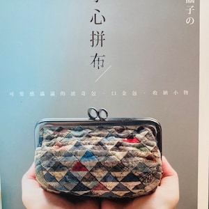 May include: A book cover with the title "Hand-Stitched Fabric" in Japanese characters. The cover features a close-up of a small, patchwork fabric purse held in two hands. The purse has a silver clasp and is made of a variety of colorful fabrics.