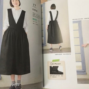 May include: A black overall dress with a deep V-neck, paired with a white blouse. The dress has a full skirt and pockets. The image is from a fashion magazine.