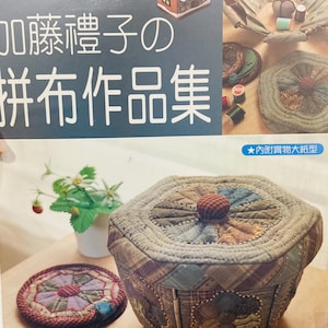 May include: A craft book cover featuring a patchwork box with a lid, a matching circular mat, and a small potted strawberry plant. The book title in Japanese characters is displayed prominently alongside images of various quilted fabric projects.